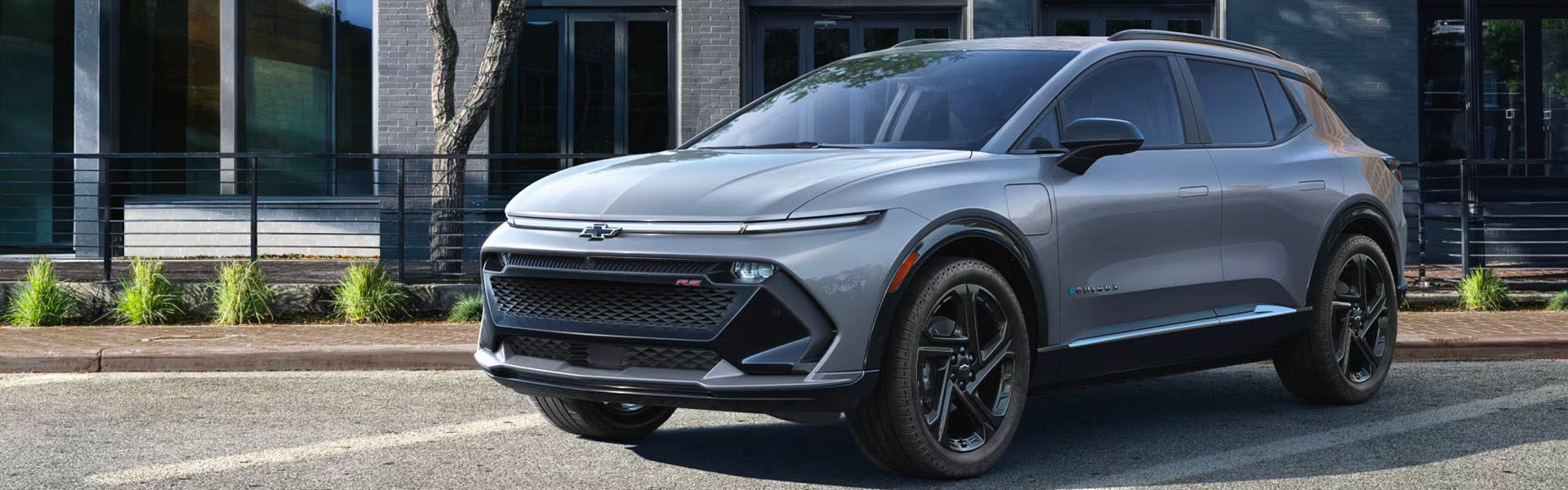 2025 Chevy Equinox EV electric SUV in gray parked on a city street, highlighting sleek aerodynamic design, closed EV grille, LED lighting, black alloy wheels, and modern Chevrolet electric crossover styling.