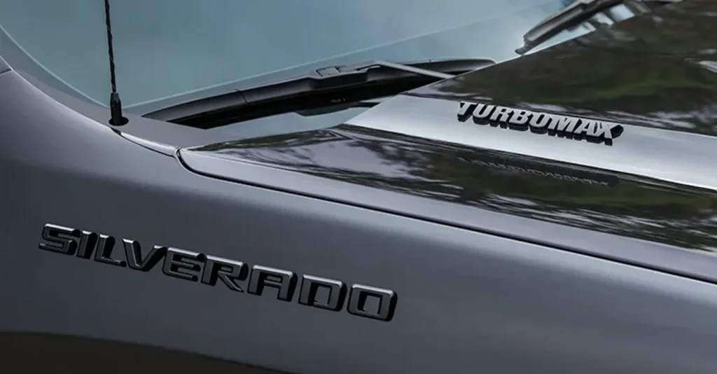 The image shows the hood and front of a 2026 Chevrolet Silverado with the "SILVERADO" badge on the left and the "TURBOMAX" emblem on the right. The sleek silver finish reflects the surroundings, adding a glossy touch to the truck's exterior design.