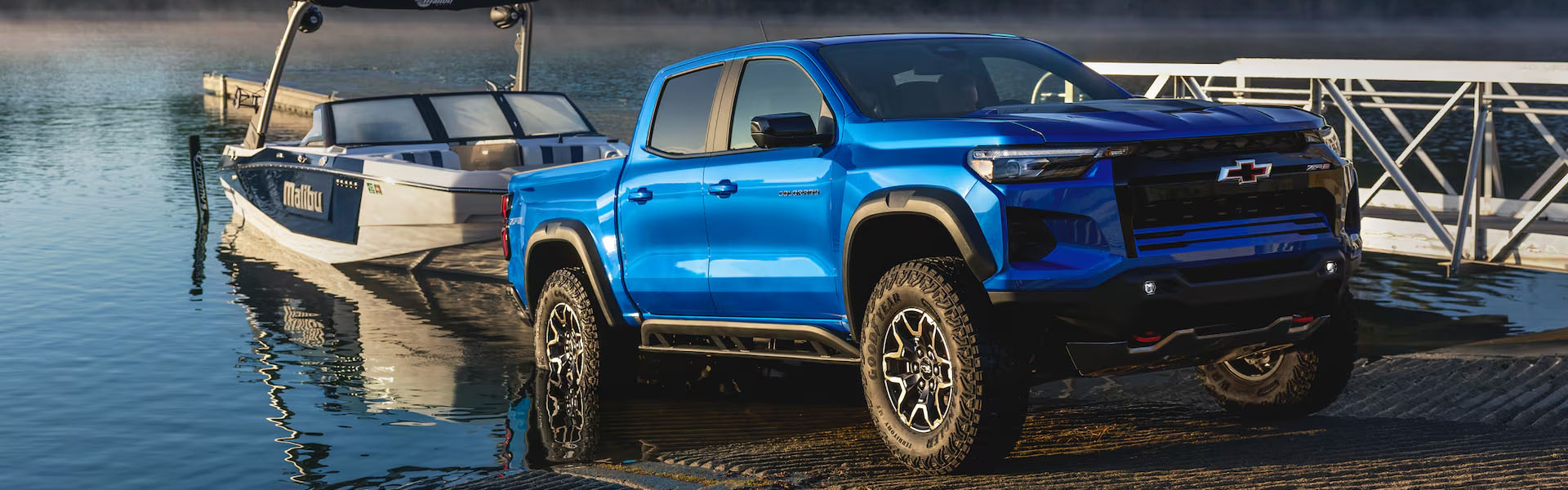 Blue 2026 Chevrolet Colorado pickup truck parked near a boat on a shimmering lake.