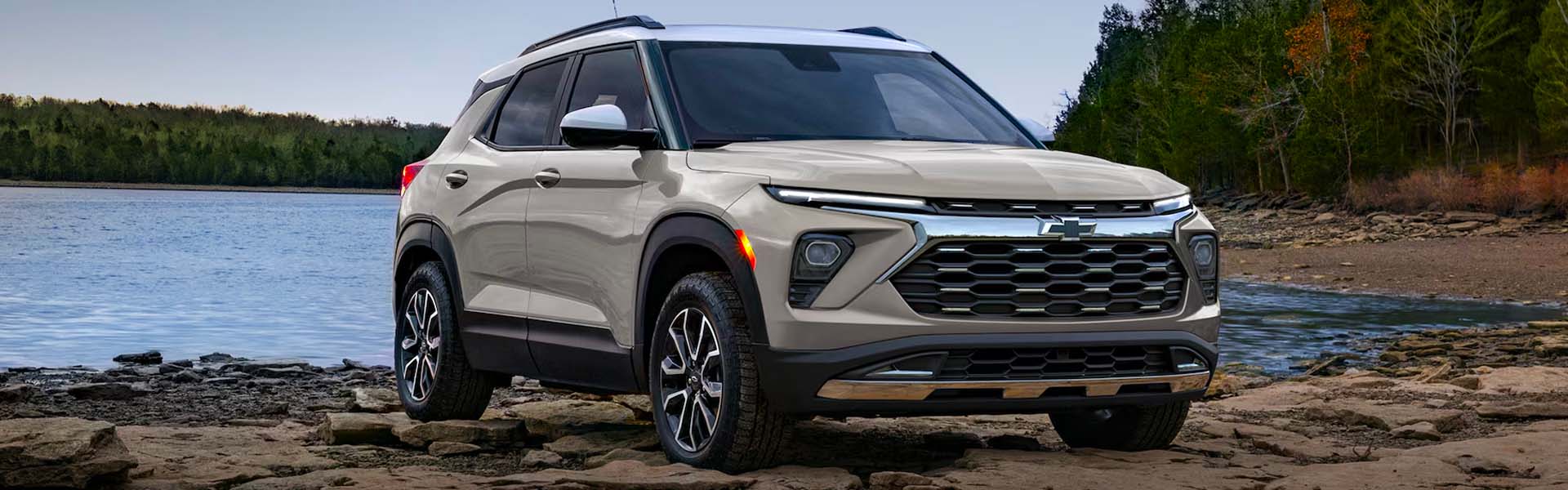 2026 Chevrolet Trailblazer parked on rocky shoreline near a calm lake, surrounded by trees.