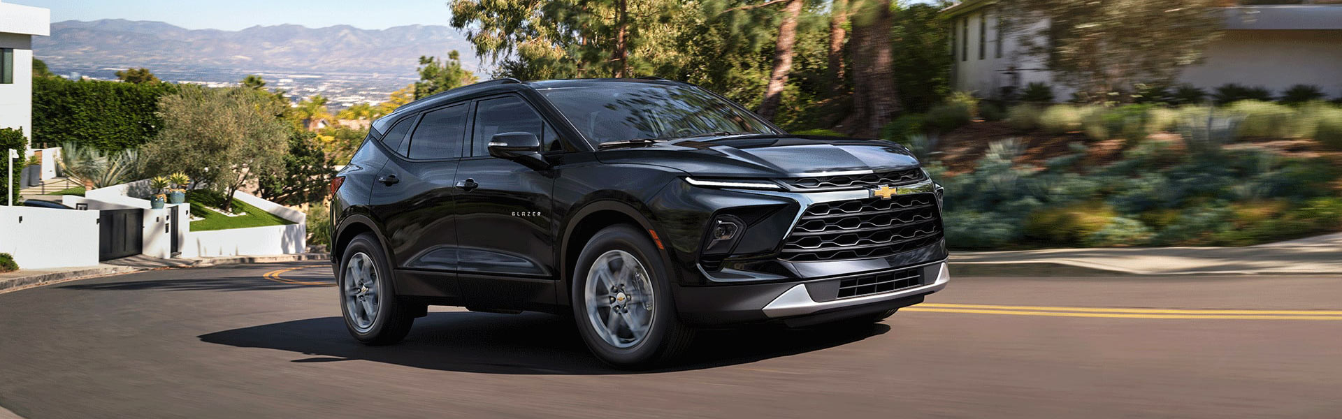 2026 Chevrolet Blazer midsize SUV in black driving on a scenic suburban road, highlighting its sporty design and bold front grille.