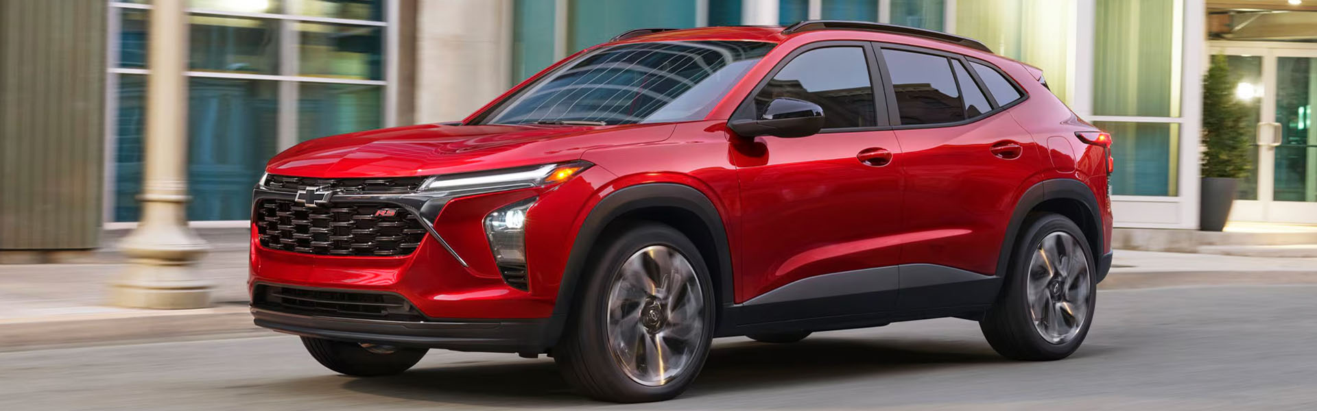 2026 Chevrolet Trax compact SUV in red driving through a downtown street, showcasing its sporty styling, bold front grille, and modern LED lighting.